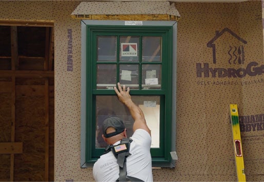 A man inserts a green-framed casement window with modified colonial grilles into a rough opening.