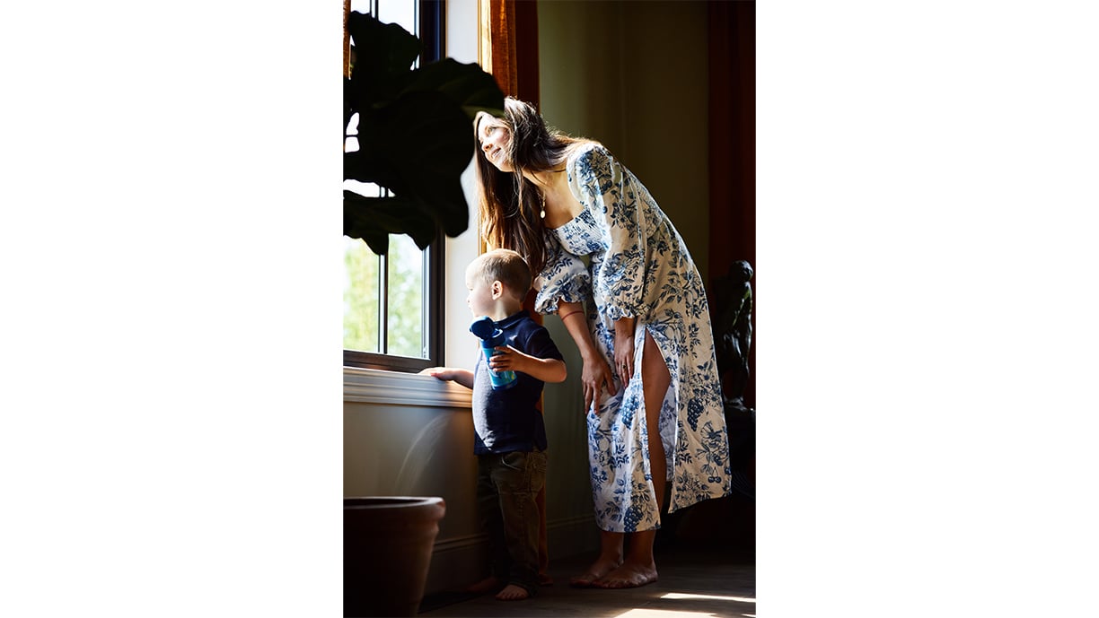 A woman wearing a white dress with blue flowers bends down next to a young boy wearing a navy-blue shirt, as they look out a window framed by ochre linen curtains and a potted plant.