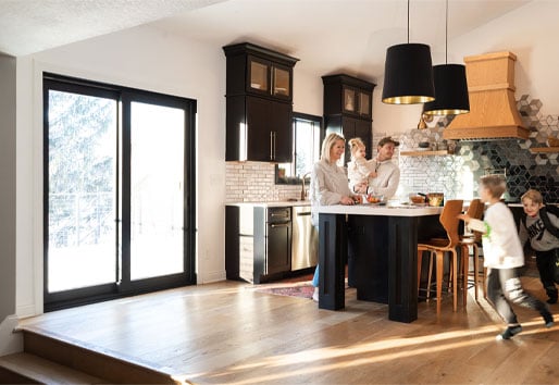 Parents laugh while their children race around a kitchen featuring black cabinetry and a black framed sliding glass door. 