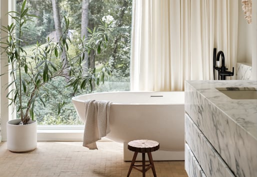 A bathroom with a large soaking tub in front of a floor-to-ceiling window that’s half covered by a white curtain and a floating vanity made of blue-veined marble.